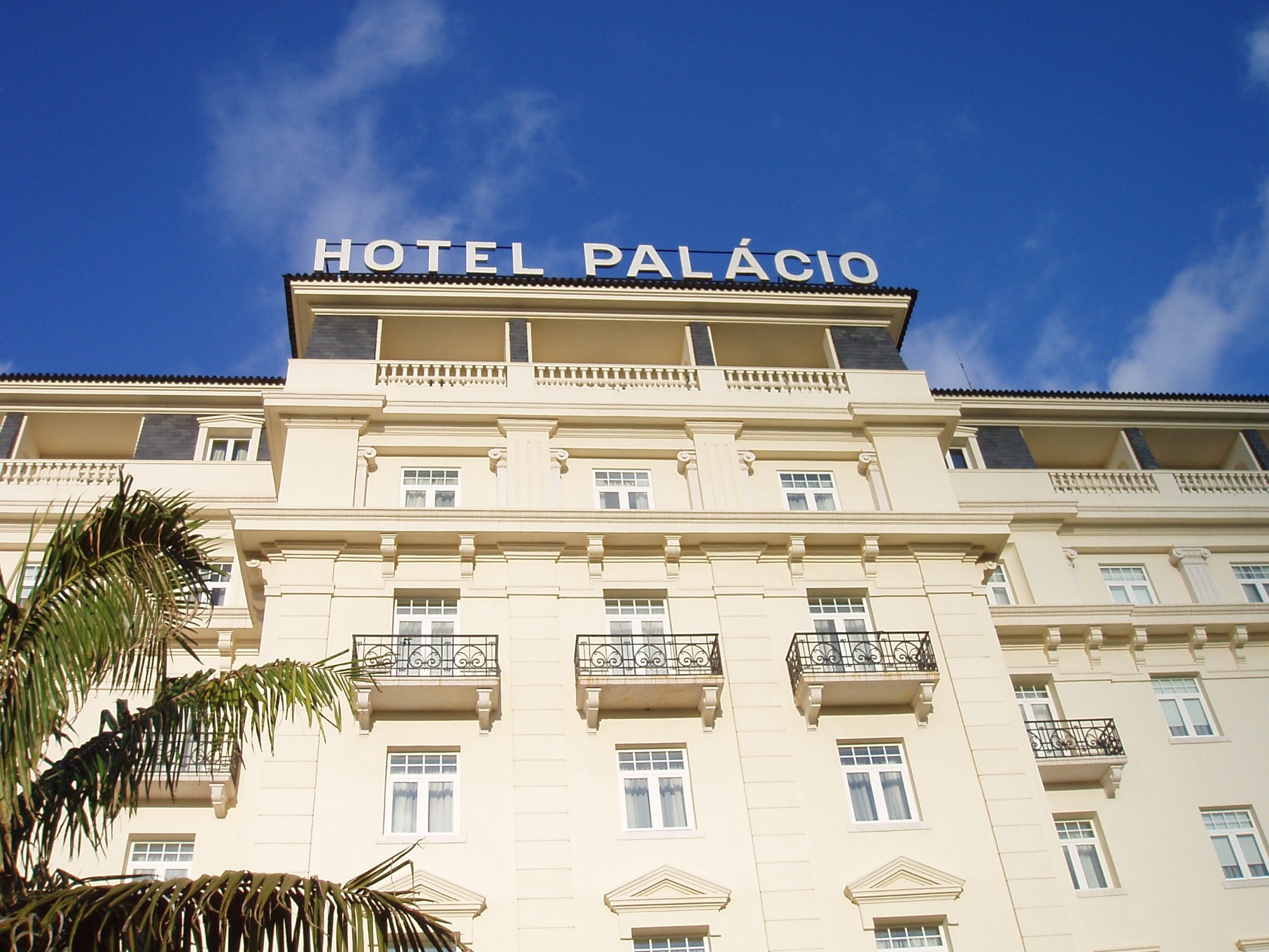 The cream façade of the Palácio Estoril Hotel with wrought-iron balconies and the words Hotel Palácio on the roofline against a blue sky