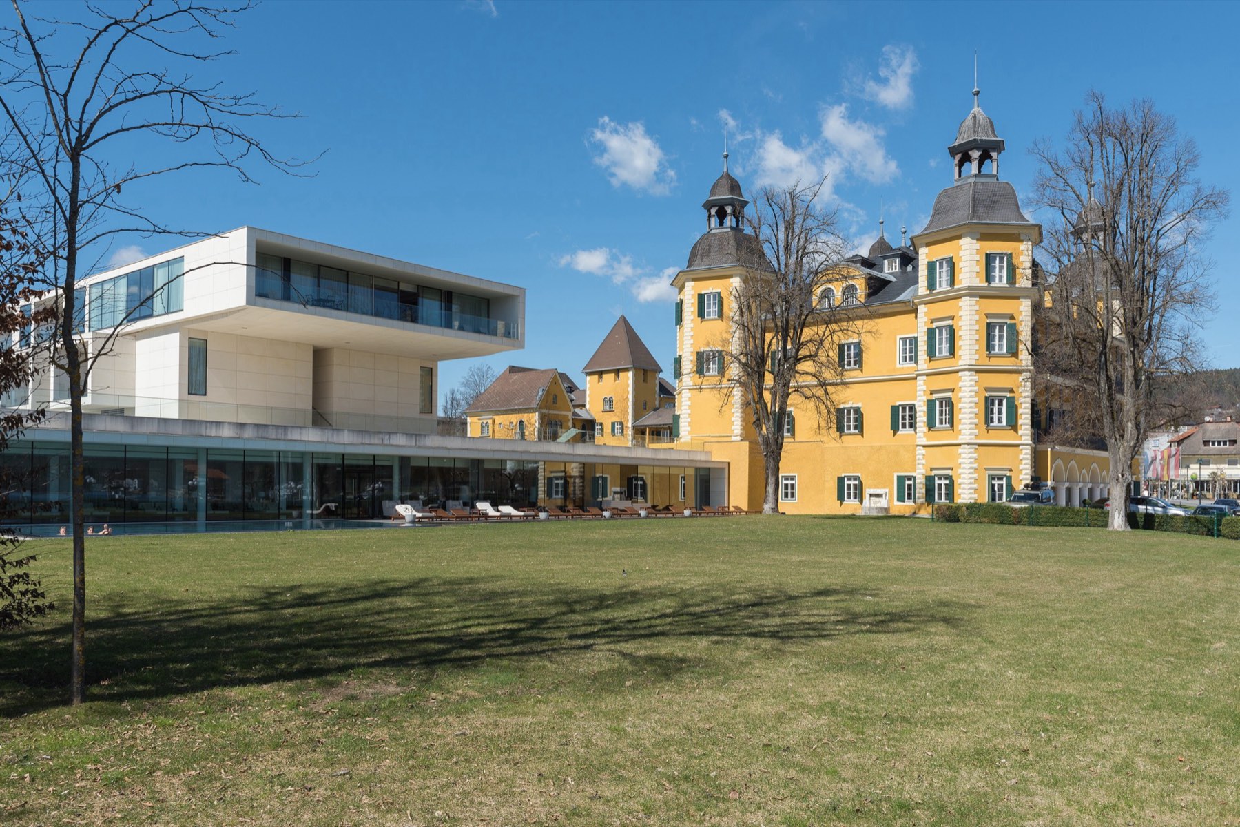 The ochre Renaissance castle of the Falkensteiner Schlosshotel Velden with its onion-domed towers, beside a modern glass-fronted wing and a lawn